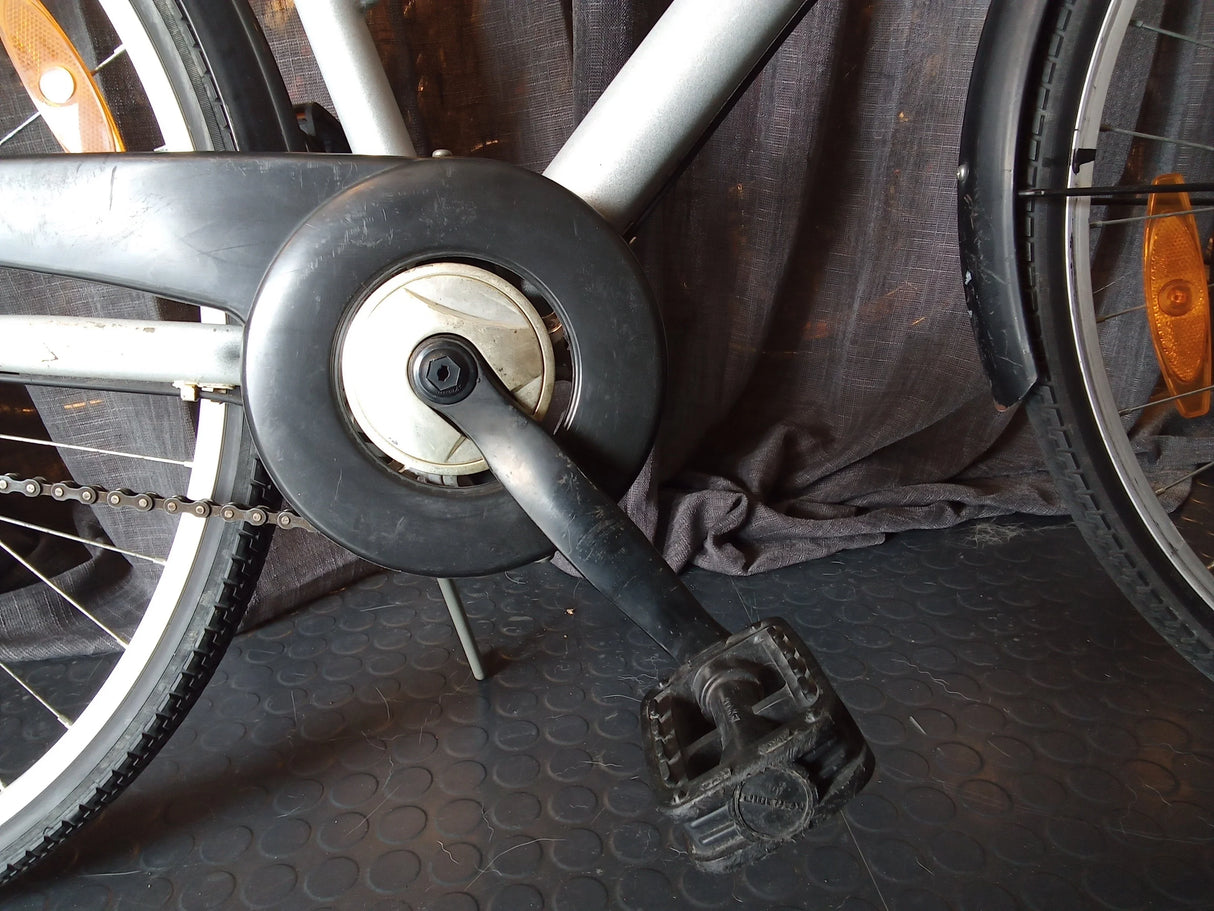 Närbild på svart cykelpedal, vevarm och kedja på silverfärgad cykelram med reflex på hjul