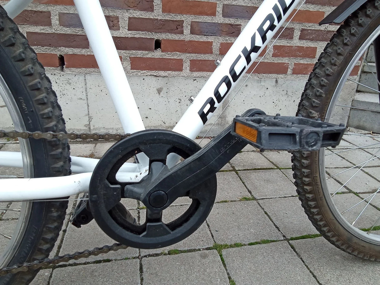 Vitt ROCKRIDER MTB med svart vevparti och pedaler med reflexer på tegelstensplatta