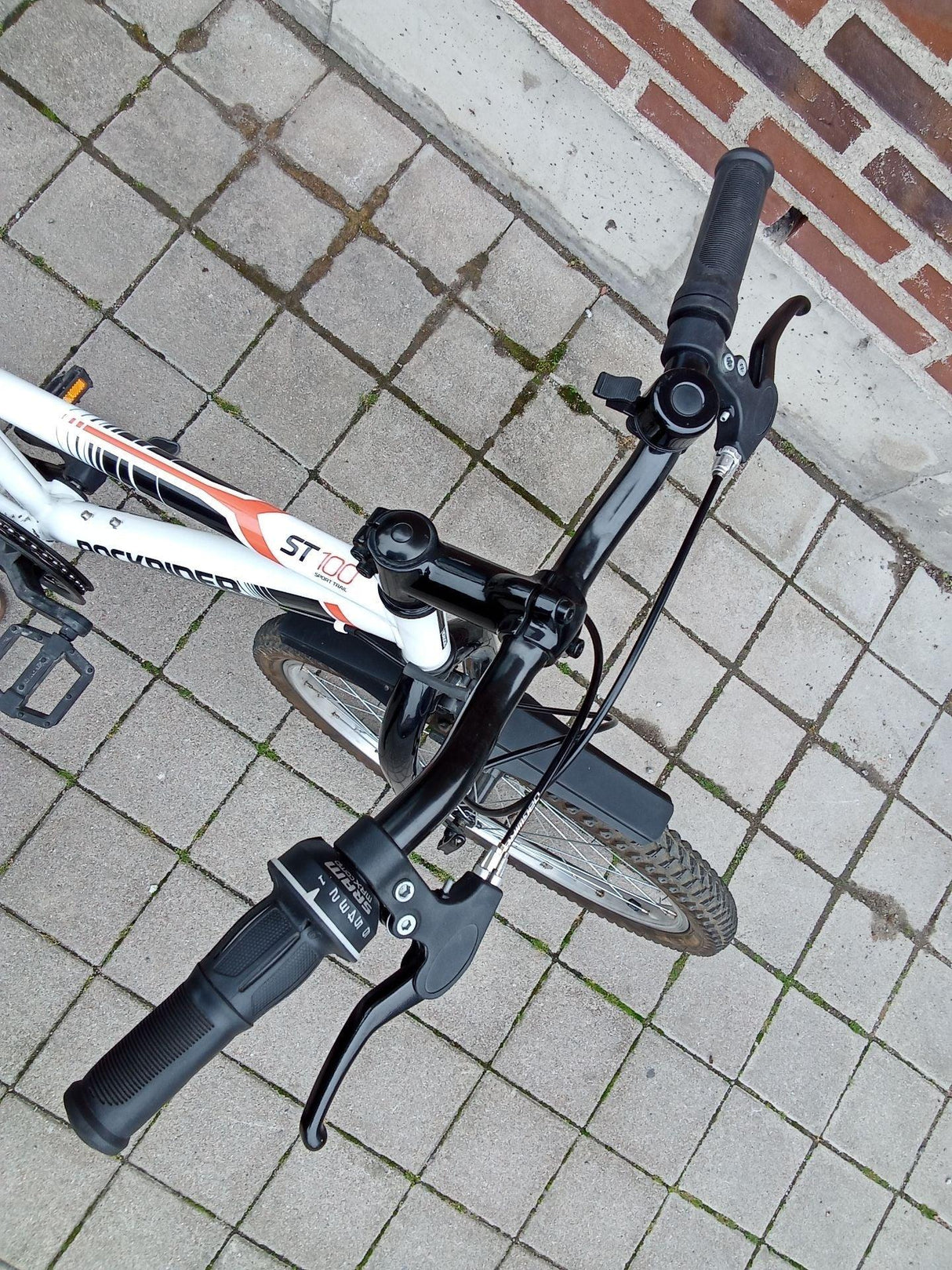 Sportig mountainbike med svart styre och vit ram märkt ST 100 på stenläggning