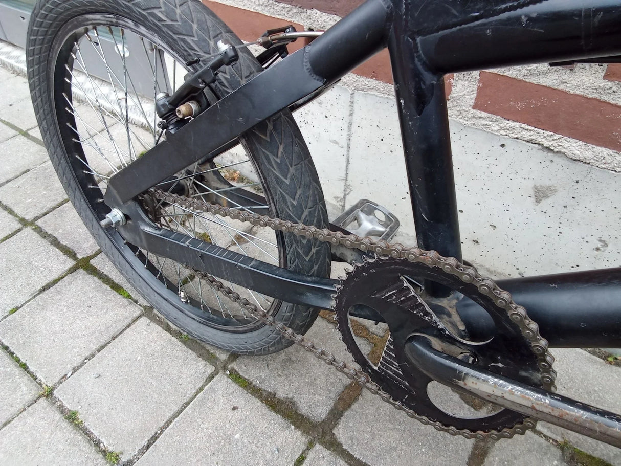 Detalj av svart BMX-cykel med kedja, vevparti och bakhjul på stenbelagd yta