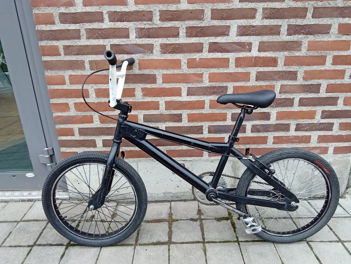 Svart BMX-cykel med vita styrehandtag och svart sadel parkerad mot tegelvägg