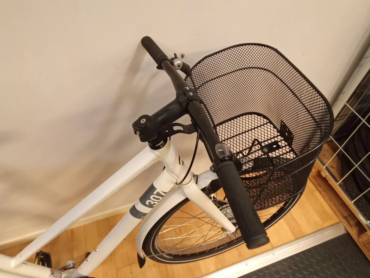 Vit cykel med svart styre och svart metallkorg fram, inomhus på trägolv