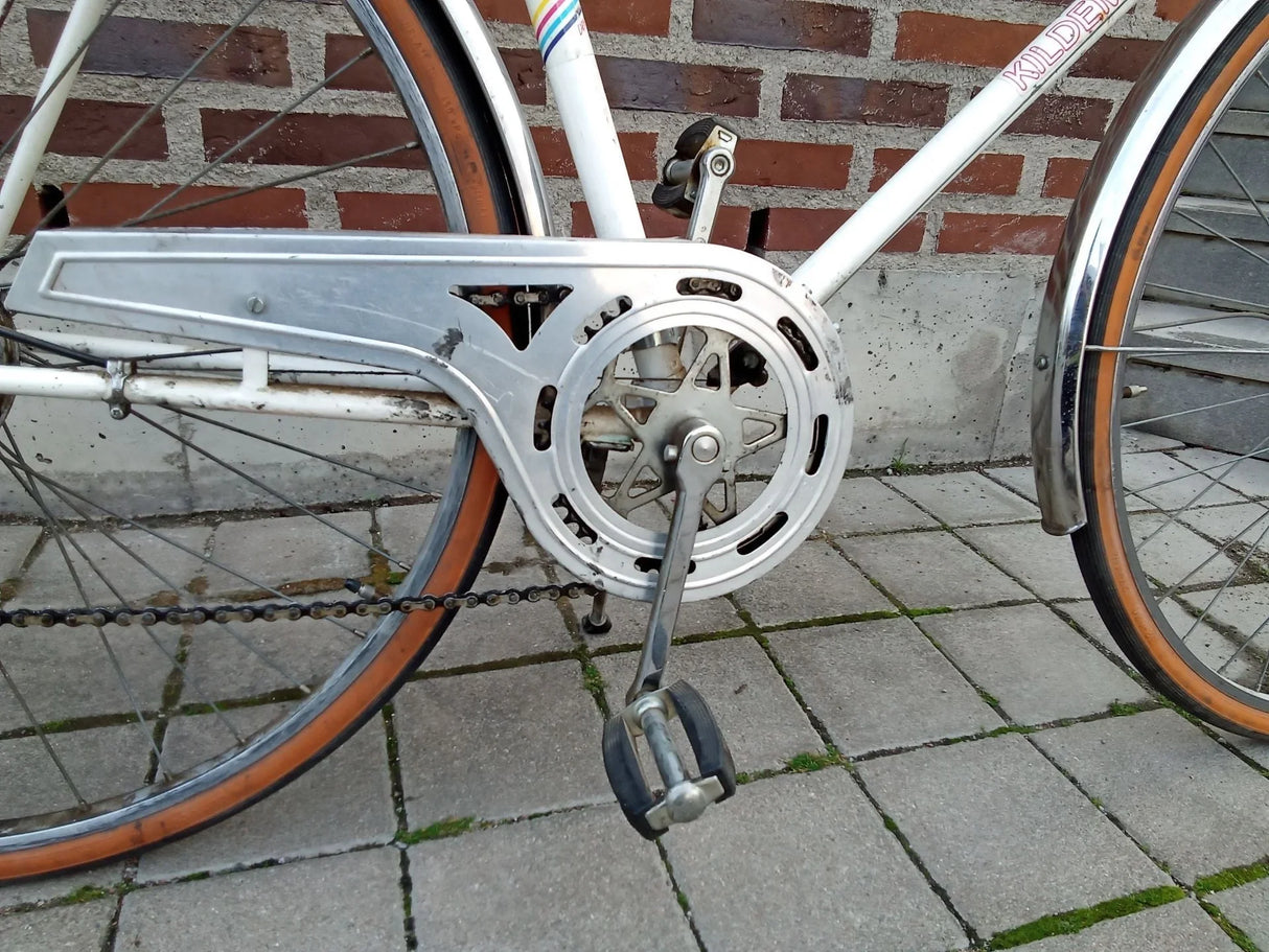 Vit vintage cykel med metallkedjeskydd och svart pedal på stenbelagd mark