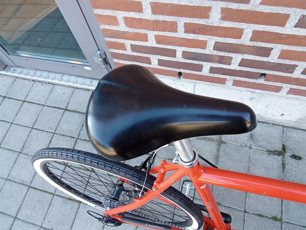 Svart cykelsadel på orange cykelram med svart däck och metallpedal