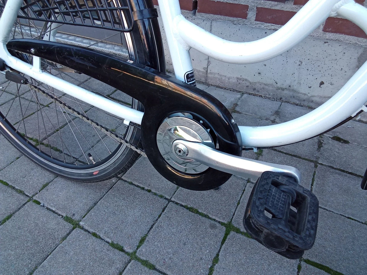 Närbild på vit cykels vevparti med svart kedjeskydd och pedal på stenlagd mark