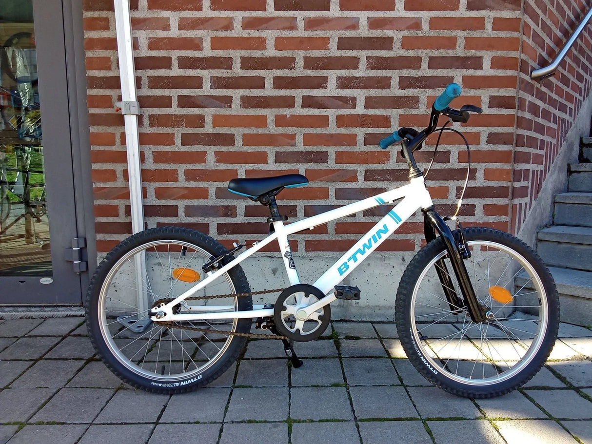 Vit B'TWIN barncykel med svart sadel och blåa handtag på stenlagd mark