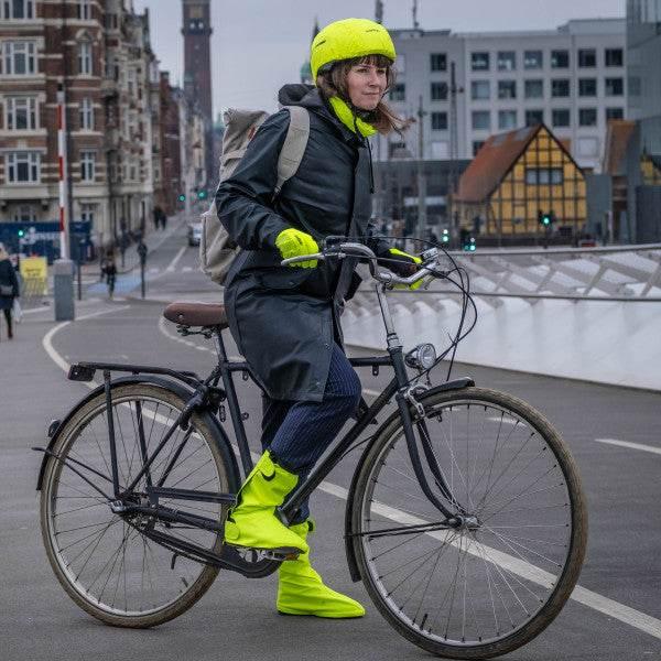 Kvinna i svart regnrock och neongula hjälm, handskar och stövlar cyklar på svart stadscykel