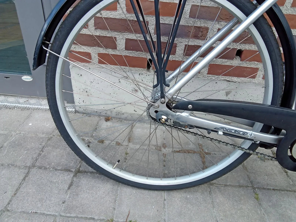 Bakre cykelhjul med svart däck, silverfärgad fälg och kedja på asfalt med tegelvägg