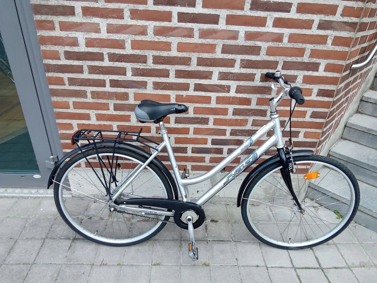 Silver citycykel med svart sadel och pakethållare på stenlagd trottoar