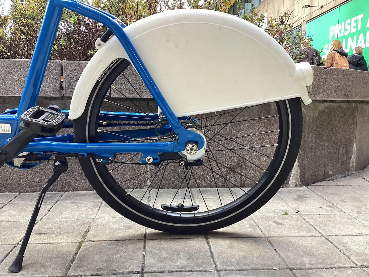 Blå cykel med vit kedjeskydd och svart bakhjul på stenlagd asfalt