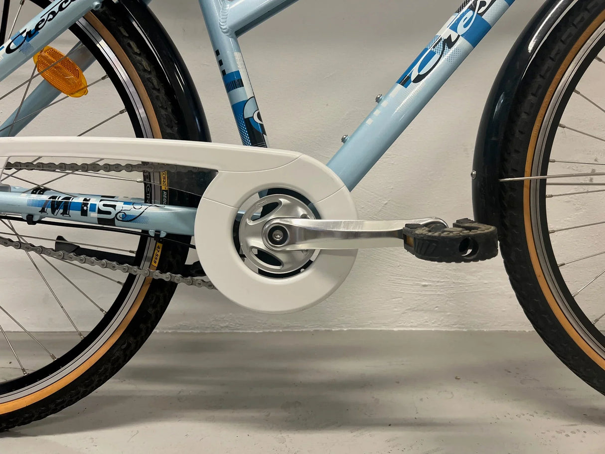 Ljusblå cykel med vitt kedjeskydd, svart däck och pedal i närbild