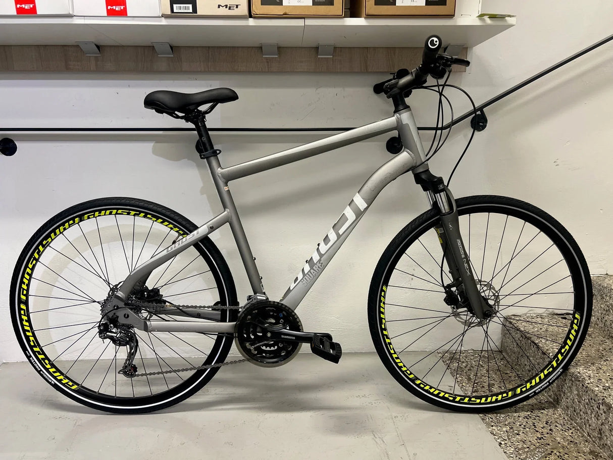 Grå Fuji hybridcykel med svart sadel och gula däcktexter mot vit vägg