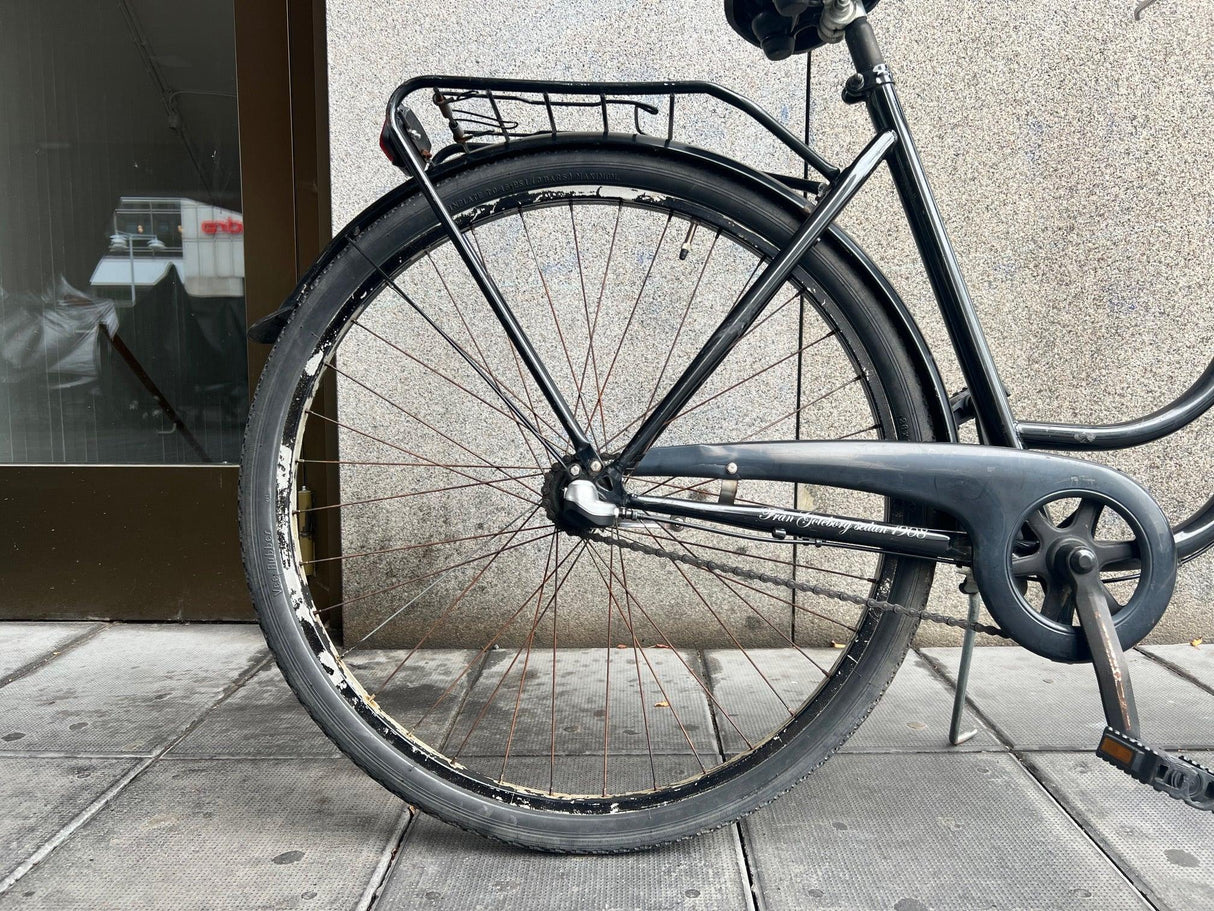 Svart citybike bakhjul med rostiga ekrar och bagagehållare på trottoar