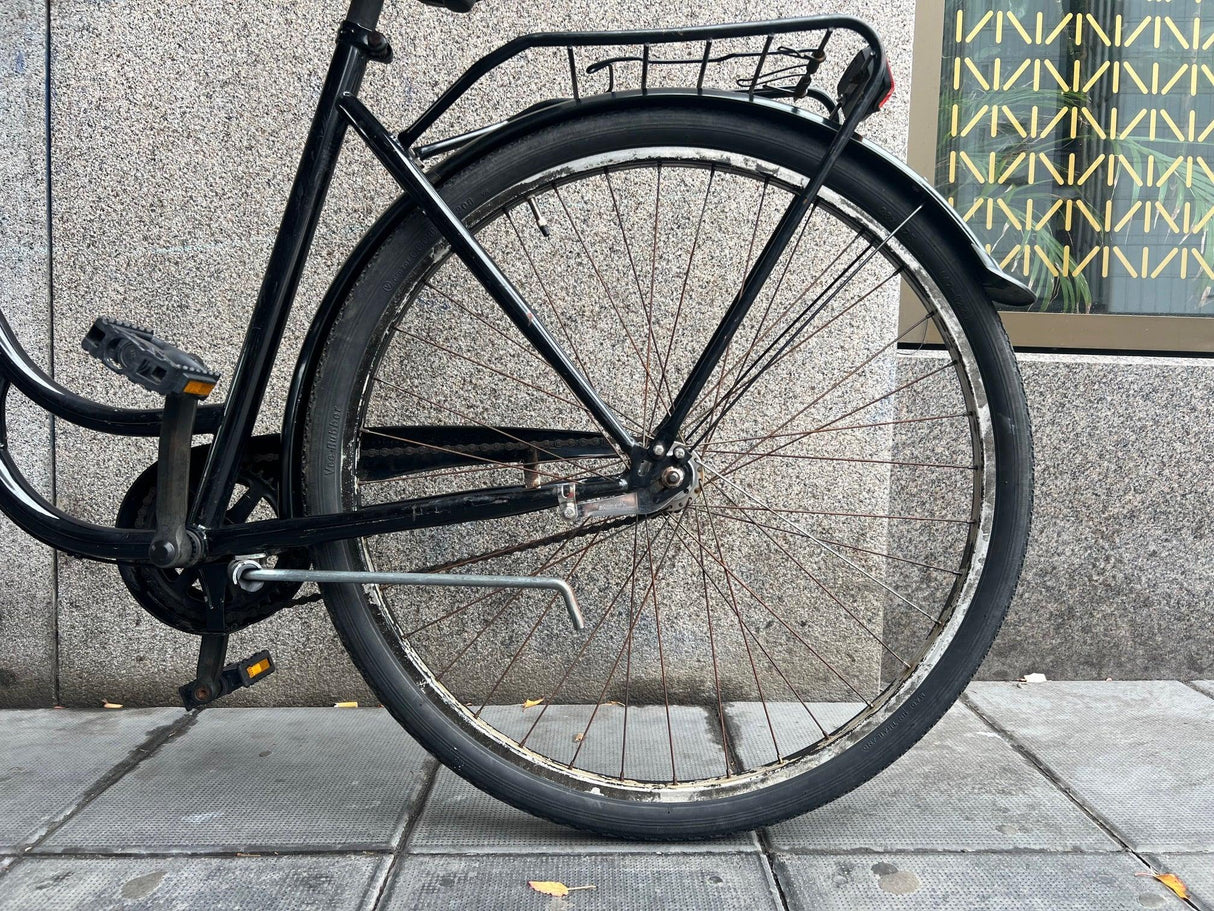 Svart stadscykel med bakhjul, pakethållare och ställning på trottoar