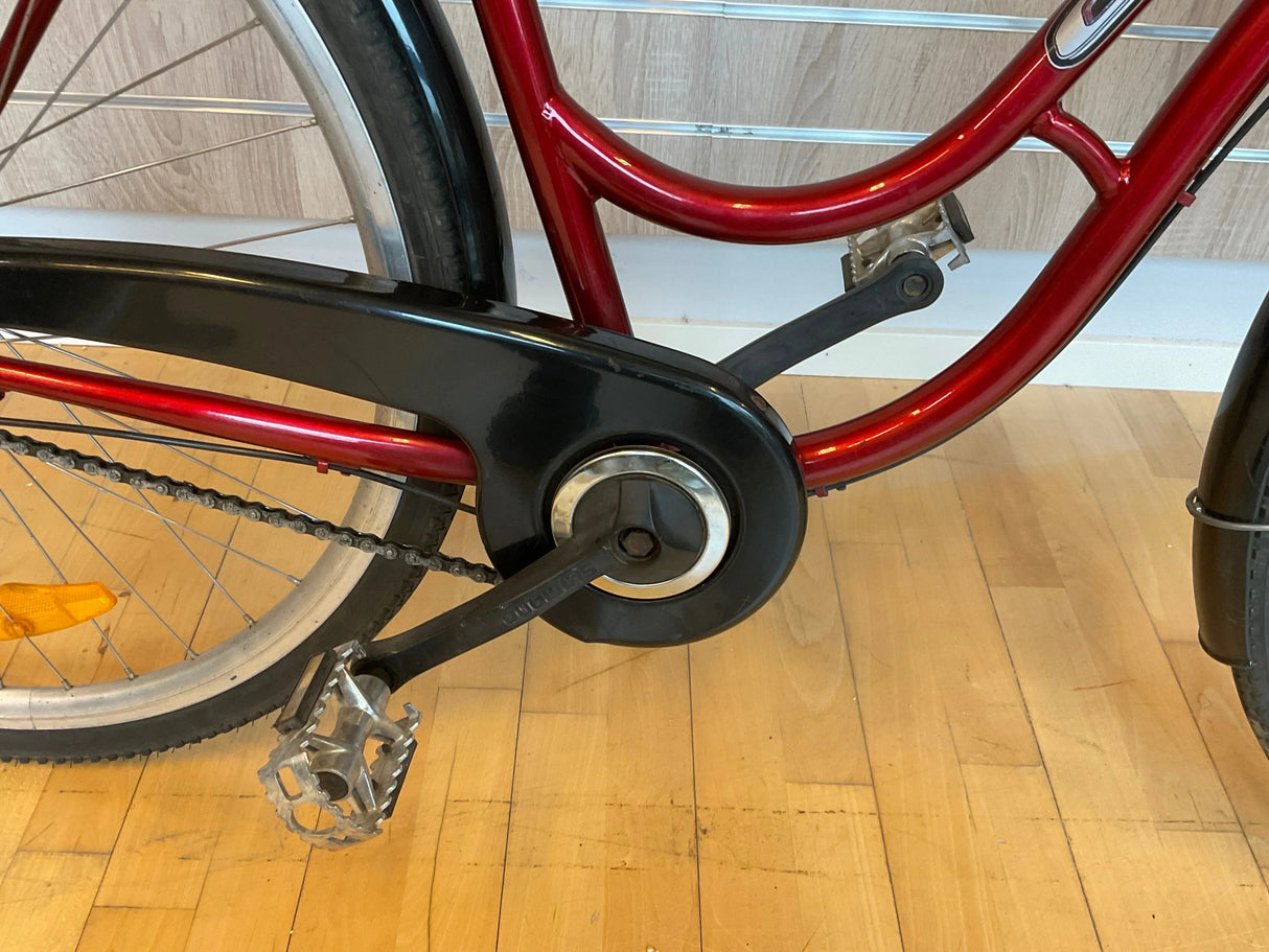 röd cykelram med svart kedjeskydd och metallpedaler på trägolv