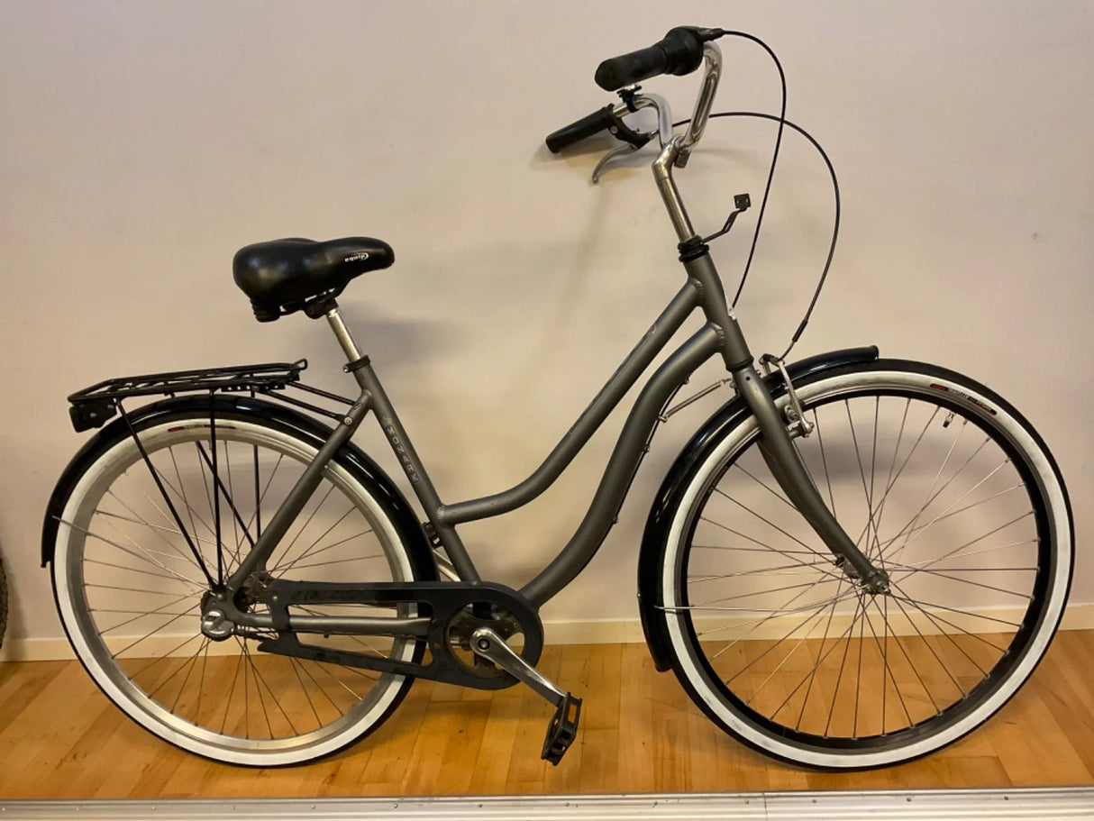 grå citybike med svart sadel, pakethållare och vita däcksidor på trägolv