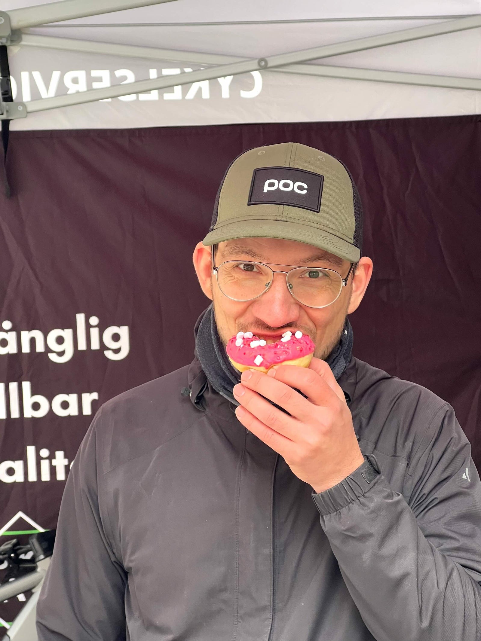 Man med glasögon och keps äter munk med rosa glasyr och strössel