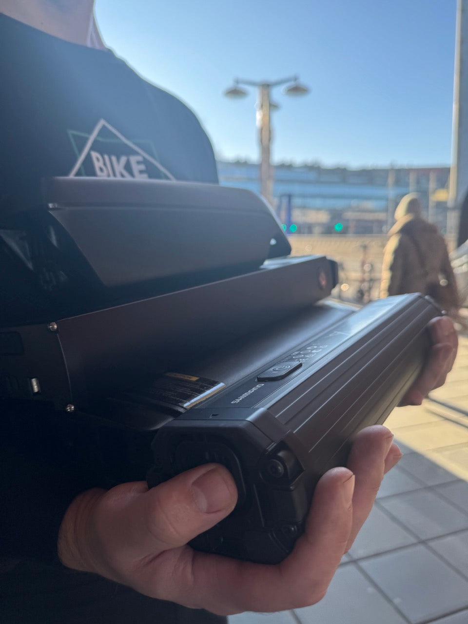 Person håller två elcykelbatterier Shimano i händerna, svart tröja med BIKE-tryck.