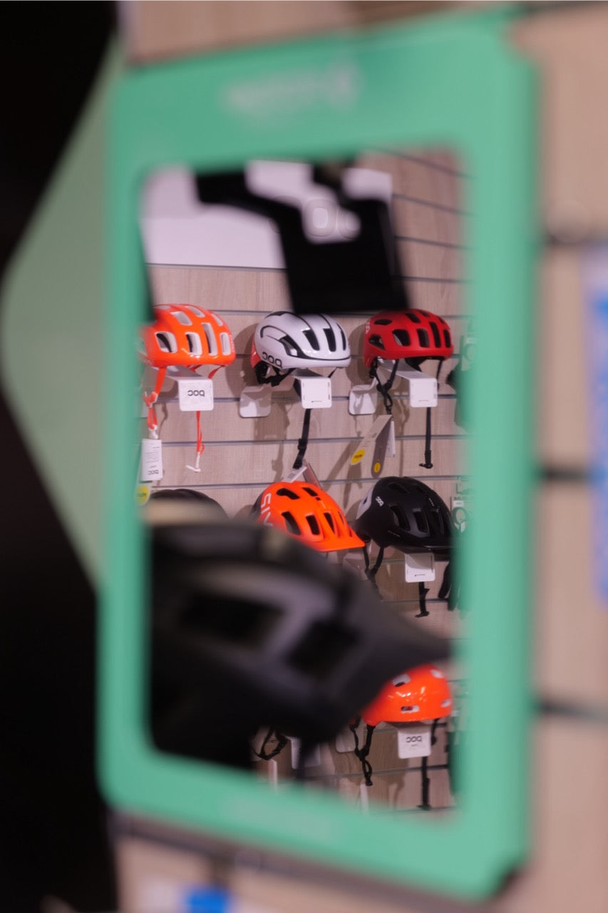 Cykelhjälmar i orange, vit, röd och svart hängande på vägg i butiksspegel