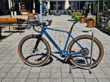 Blå Canyon gravel-cykel med breda däck, svart sadel och styre i cykelställ
