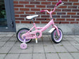 rosafärgad barncykel med stödhjul och vit sadel, märke Pollux