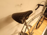Vintage grå cykel med svart slitnare sadel och raka styrstänger på trägolv