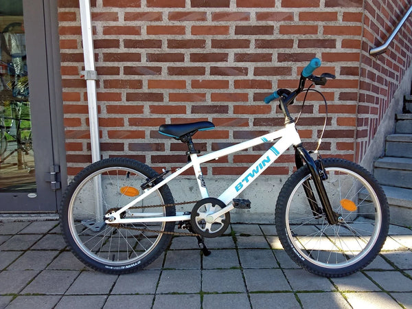 Vit B'TWIN barncykel med svart sadel och blåa handtag på stenlagd mark
