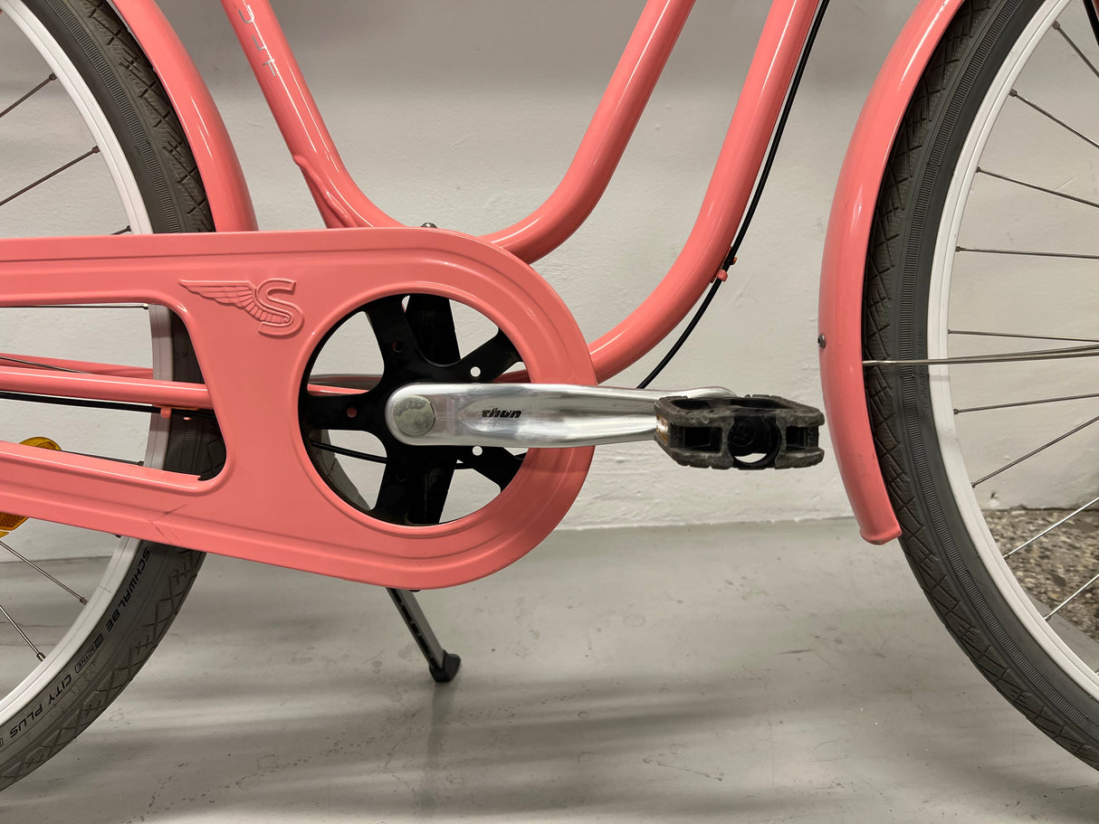 Rosafärgad cykel med kedjeskydd och pedal, detalj på vevarm och däck