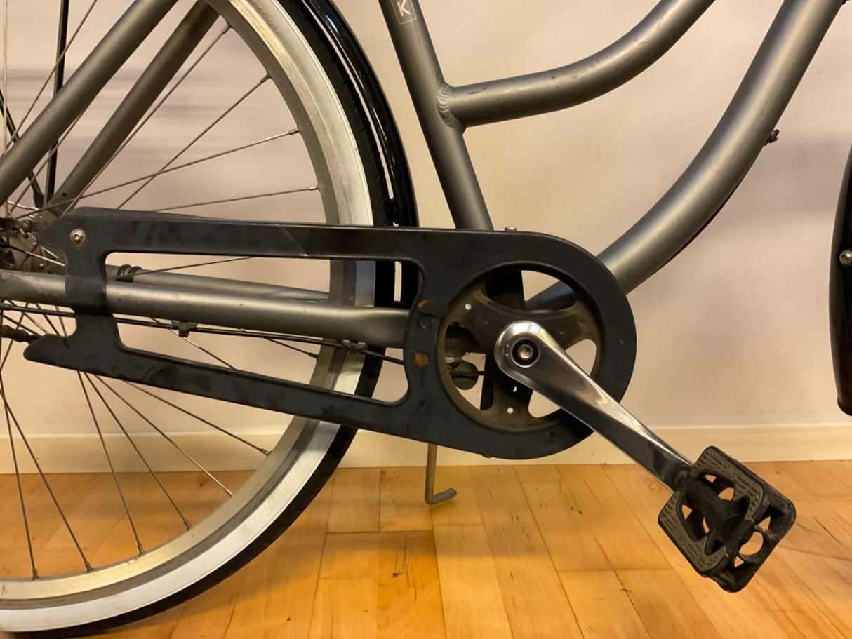 Närbild av cykelns kedjeskydd, pedal och bakhjul med vitt däck och grå ram