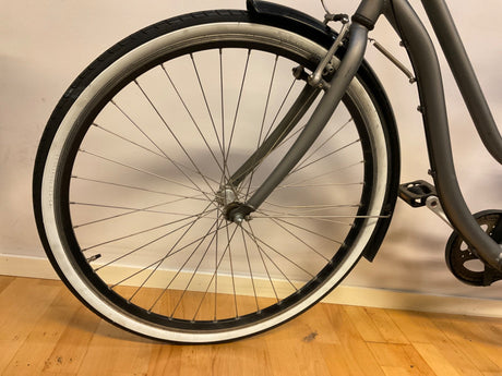 Framhjul på grå cykel med svart och vit kant på däcket, cykelpedal och skärm syns